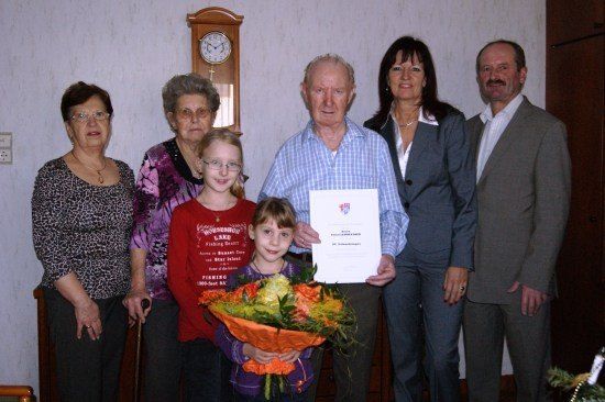 Eine Gruppe von Menschen, darunter zwei ältere Frauen, ein junges Mädchen mit Blumen und zwei Männer, stehen in einem Raum zusammen. Ein Mann hält ein Zertifikat.