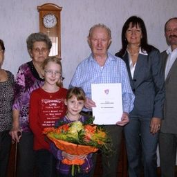Eine Gruppe von Menschen, darunter zwei ältere Frauen, ein junges Mädchen mit Blumen und zwei Männer, stehen in einem Raum zusammen. Ein Mann hält ein Zertifikat.