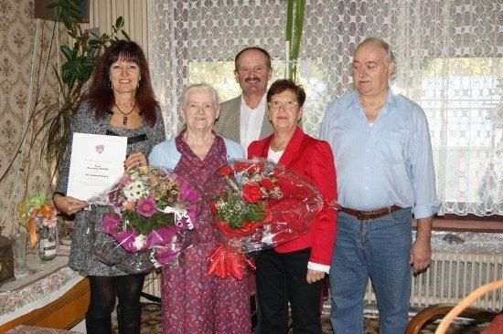 Eine Gruppe von Menschen, einschließlich einer älteren Frau, steht zusammen und hält Blumen und ein Zertifikat. Sie lächeln und posieren für ein Foto.