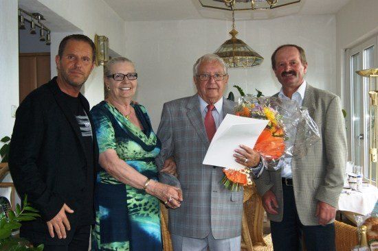 Vier Personen stehen in einem Raum, ein Mann hält einen Blumenstrauß und ein Papier und lächelt. Dahinter steht ein Tisch mit einer weißen Tischdecke.