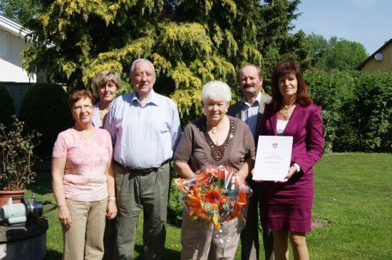 Eine Gruppe von Menschen steht in einem Garten, lächelt und posiert für ein Foto. Eine Frau hält einen Blumenstrauß und eine andere Frau hält ein Zertifikat.