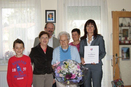 Eine Gruppe von Menschen steht in einem Raum, lächelt und posiert für ein Foto. Eine Frau hält einen Blumenstrauß und ein Zertifikat. Ein Mann steht hinter ihr und eine Frau steht neben ihm. Ein Bilderrahmen ist an der Wand montiert.