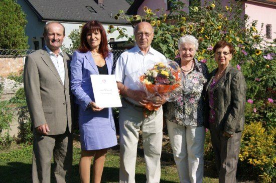 Fünf Menschen stehen in einem Garten und lächeln für ein Foto. Ein Mann hält einen Blumenstrauß und ein Zertifikat. Hinter ihnen ist ein Haus mit einem Zaun und Sonnenblumen zu sehen.