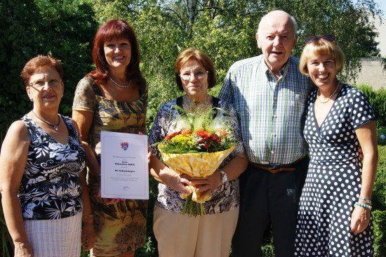 Fünf Menschen stehen in einem Garten und lächeln für ein Foto. Eine Frau in der Mitte hält ein Zertifikat und einen Blumenstrauß. Im Hintergrund sind Grünpflanzen und Bäume zu sehen.