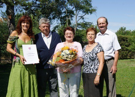 Fünf Menschen stehen lächelnd in einem Garten. Eine Frau hält einen Blumenstrauß und ein Zertifikat. Der Mann rechts trägt ein weißes Hemd, der Mann links ein blaues Sweatshirt. Hinter ihnen stehen Bäume und Büsche.