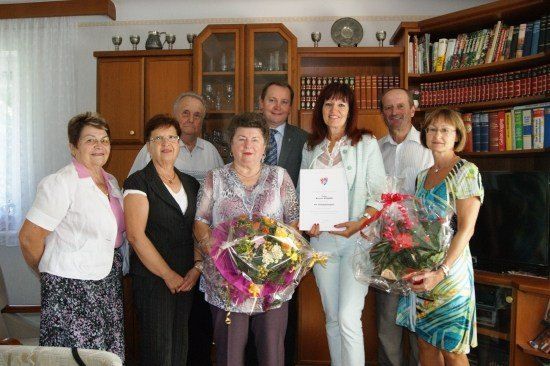 Eine Gruppe von Menschen steht in einem Raum und hält Blumen und ein Zertifikat. Eine Frau in der Mitte hält das Zertifikat und Blumen. Hinter ihnen sind Holzschränke mit Büchern und Dekorationsartikeln.