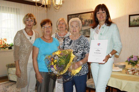 Fünf Frauen stehen in einem Raum, eine hält einen Blumenstrauß und eine andere ein Zertifikat. Sie lächeln und scheinen ein Ereignis zu feiern.