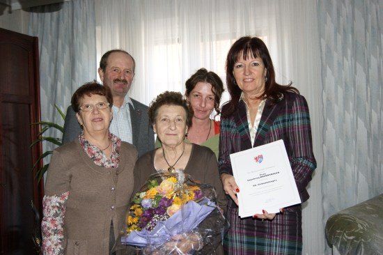 Eine Gruppe von Menschen, darunter eine ältere Frau mit Blumen und einem Zertifikat, steht in einem Raum mit einem Fenster und Vorhängen im Hintergrund.