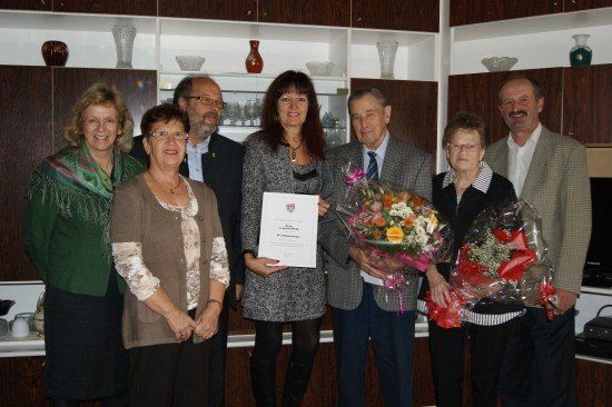 Eine Gruppe von Menschen steht lächelnd in einem Raum. Eine Frau hält ein Zertifikat und Blumen. Hinter ihnen befinden sich Regale mit verschiedenen Gegenständen.