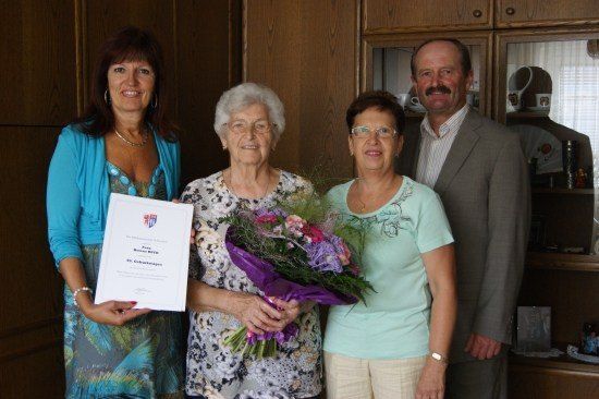 Eine Frau mit einem Zertifikat und Blumen steht zwischen zwei anderen Frauen, alle lächeln, mit einem Mann im Anzug hinter ihnen.