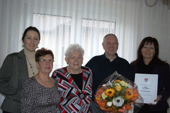 Fünf Personen stehen vor einem weißen Vorhang. Die Frau in der Mitte hält einen Blumenstrauß und ein Zertifikat. Die Frau rechts hält ein Zertifikat. Die Frau links trägt eine Halskette. Der Mann in der Mitte trägt eine Brille.