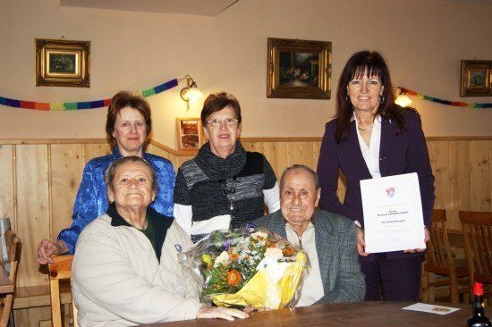 Eine Gruppe älterer Menschen versammelt sich um einen Tisch, wobei ein Mann einen Blumenstrauß und ein Zertifikat hält. Hinter ihnen steht eine Frau, die ein Zertifikat hält und lächelt. Der Raum ist mit gerahmten Bildern und bunten Wimpelketten dekoriert.