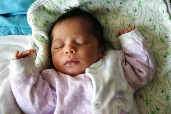 Ein Neugeborenes schläft friedlich auf einem Bett und trägt ein rosa-weißes Pünktchen-Outfit mit Schmetterlingsdesign. Das Baby hat die Hände leicht erhoben und liegt auf einer Decke mit grünen Bären-Designs.