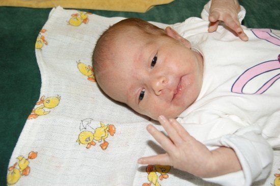 Ein lächelndes Baby liegt auf einer weißen Decke mit einem gelben Entenmuster und trägt ein weißes Shirt mit einem rosa Design.