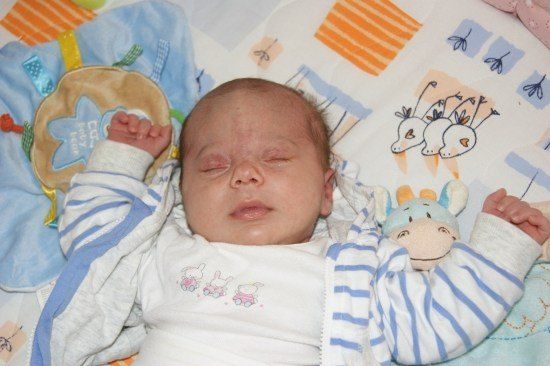 Ein Baby schläft auf einem Bett mit einer Decke und einem Kuscheltier. Das Baby trägt ein weiß-blau gestreiftes Outfit.