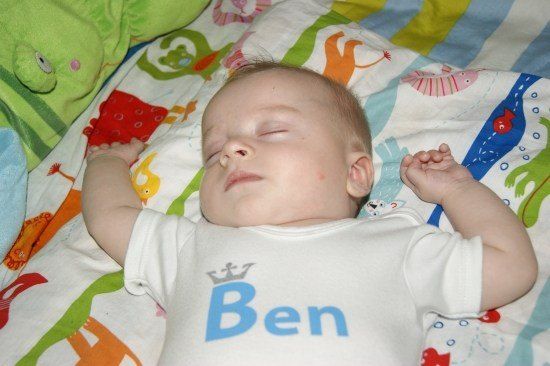 Ein Babyjunge namens Ben schläft friedlich auf einer bunten Decke mit Tierdruck, trägt ein weißes T-Shirt mit seinem Namen.