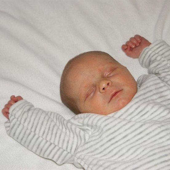 Ein Neugeborenes schläft auf einer weißen Decke und trägt einen weißen und grau gestreiften Strampler. Die Hände des Babys sind leicht gekrümmt.