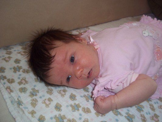 Ein Babymädchen liegt auf einem Bett mit einem rosa Deckchen und Bären darauf, trägt ein rosa Kleid mit Rüschen.