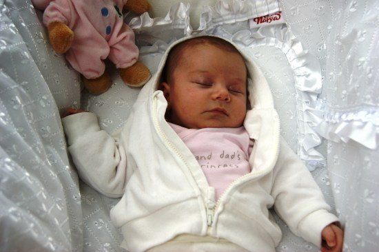 Ein Baby in einer weißen Kapuzenjacke schläft friedlich in einem Kinderbett, mit einem rosa Plüschtier in der Nähe.