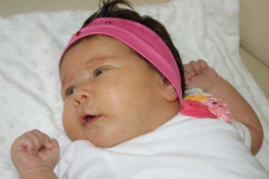 Ein Babymädchen mit einem rosa Stirnband liegt auf einer weißen Decke und lächelt mit leicht geöffnetem Mund.