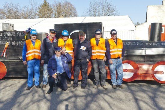 Eine Gruppe von Männern in Sicherheitswesten und Mützen posiert vor einer großen, farbenfrohen zugähnlichen Struktur.