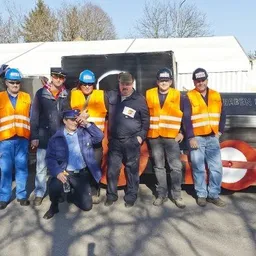 Eine Gruppe von Männern in Sicherheitswesten und Mützen posiert vor einer großen, farbenfrohen zugähnlichen Struktur.