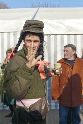Eine Person in einem grünen Kostüm mit Gesichtsbemalung und Hut steht und hält einen Becher, während eine andere Person in einer orangefarbenen Jacke zusieht.