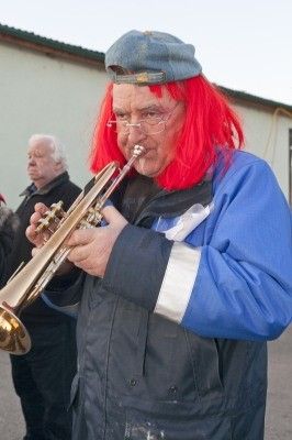 Ein älterer Mann mit roten Haaren und Brille spielt eine Trompete im Freien, trägt einen blauen Hut und eine Jacke.