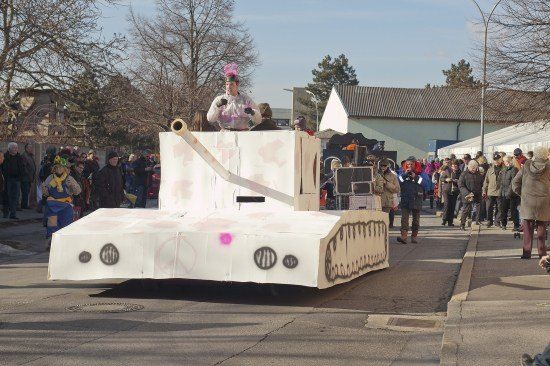 Ein Panzer-förmiger Wagen fährt die Straße entlang, während Menschen in Kostümen zusehen.