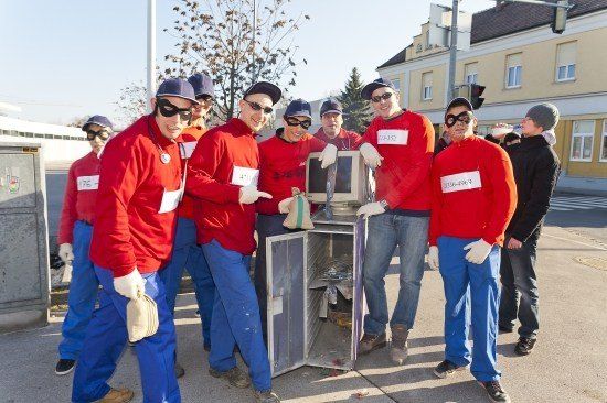 Eine Gruppe von Menschen in roten Overalls und Masken steht um einen kaputten Computer auf der Straße.