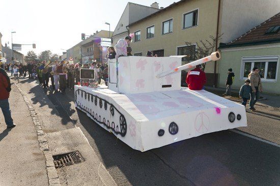Ein Paradewagen in Form eines Panzers mit Friedenssymbolen, mit Menschen, die auf der Straße gehen und Gebäude im Hintergrund.