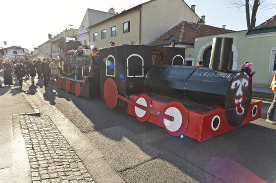 Ein dekorierter zugförmiger Umzugswagen fährt die Straße entlang, während Menschen vom Bürgersteig zuschauen.