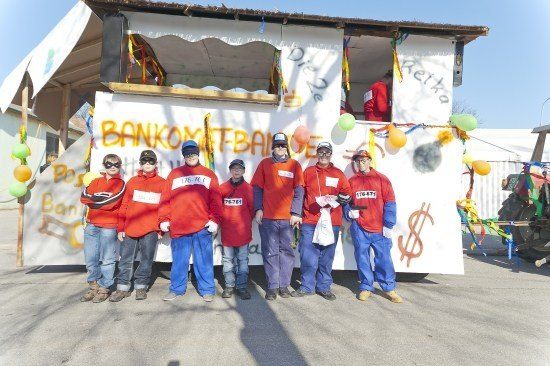 Eine Gruppe von Menschen in roten und blauen Kostümen posiert für ein Foto vor einem geschmückten Wagen mit weißem Hintergrund und Dollarzeichen.