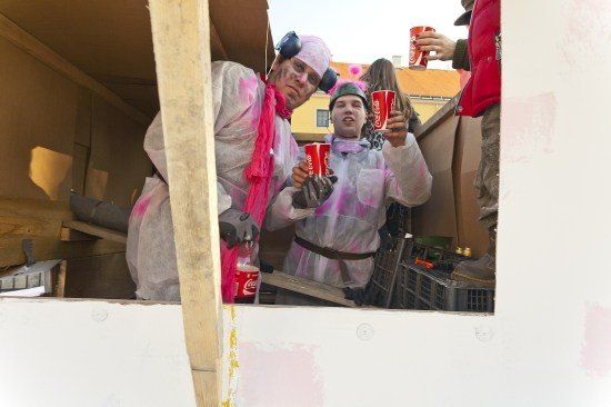 Zwei Personen in Schutzanzügen und Kopfhörern halten rote Coca-Cola-Becher in einer Pappschachtel, während eine dritte Person in einem roten Jackett draußen einen Becher hält.