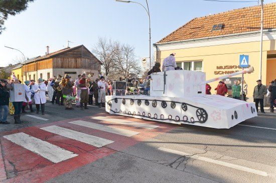 Ein Umzug mit einem großen Panzerwagen, Leuten in Kostümen und einem Gebäude mit dem Schild 'MUSIK-SCHWARZ'.