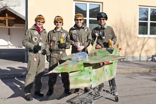 Vier Personen in Pilotenuniformen stehen neben einem Wagen mit einem Pappjet. Sie halten Dosen in den Händen und scheinen für ein Foto zu posieren.