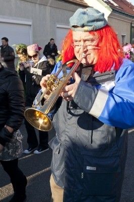 Ein Mann mit roten Haaren und einem blauen Hut spielt eine Trompete bei einer Straßenveranstaltung, mit anderen Leuten im Hintergrund.
