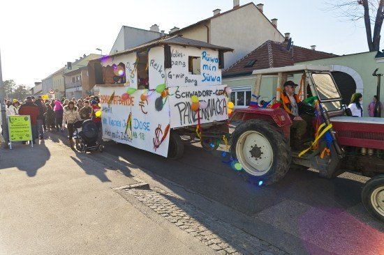Ein Traktor mit einem geschmückten Anhänger führt eine Parade eine Straße hinunter, mit Zuschauern und Gebäuden im Hintergrund.