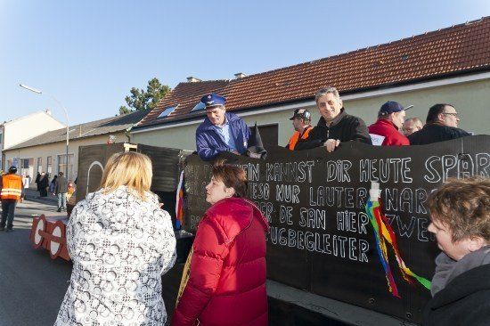 Eine Gruppe von Menschen versammelt sich um ein Fahrzeug mit einem Banner, auf dem 'In kannst dir heute Sperr' steht. Ein Mann in einem blauen Hut steht auf dem Fahrzeug.