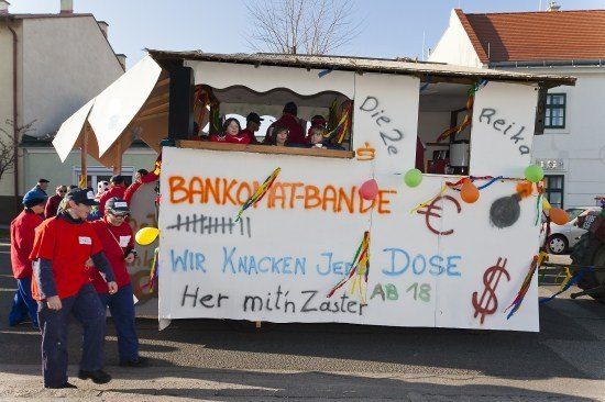 Ein geschmückter Wagen mit Menschen im Inneren, der ein Banner mit bunten Graffiti und Luftballons zeigt. Der Wagen heißt 'Bankomat-Bande' und nimmt an einem Umzug teil.