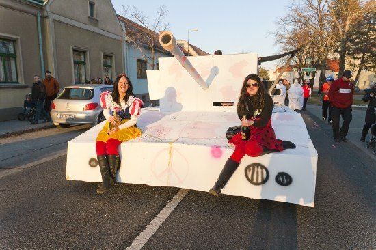 Zwei Frauen in Kostümen sitzen auf einem weißen Panzerwagen während einer Parade, mit Zuschauern und Gebäuden im Hintergrund.