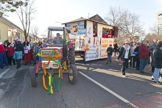 Ein dekorierter Traktor führt eine Parade an, hinter ihm gehen Menschen in Kostümen auf einer Straße, mit Bäumen und Gebäuden im Hintergrund.