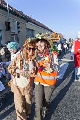Zwei Frauen in Kostümen gehen während eines Umzugs die Straße entlang. Eine trägt ein braunes Kostüm mit einer Pflanze auf ihrem Hut, und die andere ist in einer orangefarbenen Warnweste.