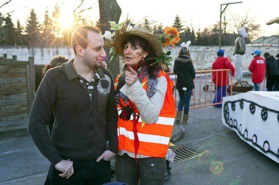 Ein Mann und eine Frau in einem Strohhut mit Blumen stehen vor einem Zaun, mit sonnenbeschienenem Hintergrund und Menschen in der Ferne.