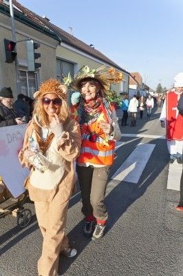 Zwei Frauen in Kostümen gehen während einer Parade die Straße entlang, eine trägt ein Löwenkostüm und die andere ein Erntearbeiter-Outfit. Hinter ihnen geht eine Menschenmenge, und Gebäude säumen die Straße.