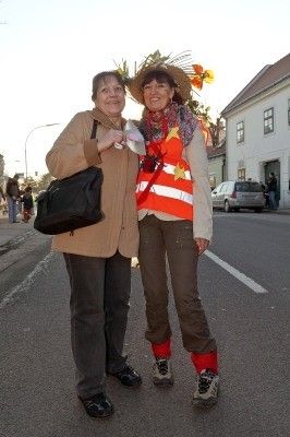 Zwei Frauen stehen an einer Straßenecke, eine trägt eine Warnweste und einen Hut mit Blumen, die andere eine Handtasche. Hinter ihnen sind ein Gebäude und ein Auto zu sehen.