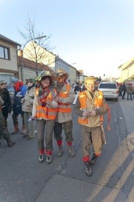 Drei Personen in orangefarbenen Westen und Mützen gehen eine Straße entlang, mit mehreren Personen im Hintergrund.