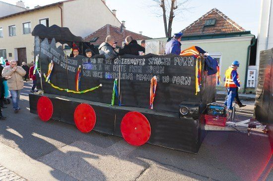 Ein dekorierter zugförmiger Wagen mit Menschen im Inneren, der sich während einer Parade durch eine Straße bewegt. Gebäude und Menschen sind im Hintergrund zu sehen.