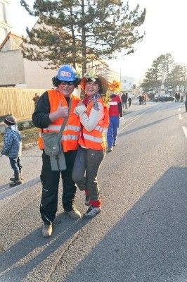 Zwei Personen in orangenen Westen und Hüten stehen auf einer Straße mit einem Kind und anderen im Hintergrund.