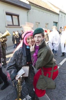 Zwei Personen in Kostümen, eine mit rosa Haaren und einer Trompete, die andere in einem grünen Barett und roten Hosen, posieren für ein Foto auf einer Straße mit einer Menge und Gebäuden im Hintergrund.